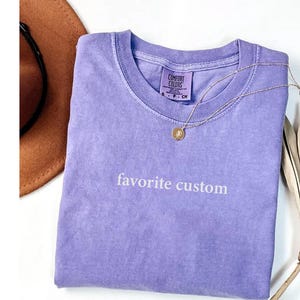 K&ouml;nnte beinhalten: Ein lavendelfarbenes Comfort Colors T-Shirt mit dem Schriftzug "favorite custom" in wei&szlig;er Schrift. Eine goldene Halskette mit Anh&auml;nger liegt auf dem Shirt. Ein brauner Hut ist teilweise in der oberen linken Ecke zu sehen.