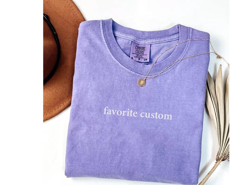 K&ouml;nnte beinhalten: Ein lavendelfarbenes Comfort Colors T-Shirt mit dem Schriftzug "favorite custom" in wei&szlig;er Schrift. Eine goldene Halskette mit Anh&auml;nger liegt auf dem Shirt. Ein brauner Hut ist teilweise in der oberen linken Ecke zu sehen.