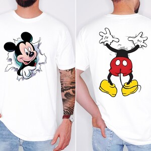 May include: A white t-shirt featuring a graphic of Mickey Mouse on the front, shown emerging from a torn paper effect. The back of the shirt displays the lower half of Mickey Mouse, including red shorts, yellow shoes, and white gloved hands.