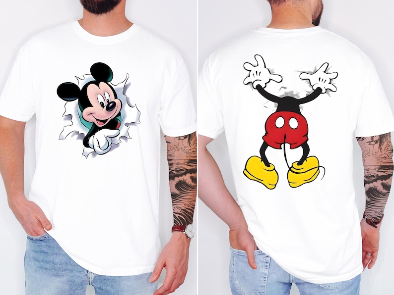 May include: A white t-shirt featuring a graphic of Mickey Mouse on the front, shown emerging from a torn paper effect. The back of the shirt displays the lower half of Mickey Mouse, including red shorts, yellow shoes, and white gloved hands.