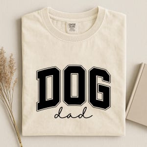 May include: Cream-colored t-shirt with the words "DOG dad" in bold black lettering. The word "DOG" is in a collegiate-style font, and "dad" is written in a cursive script. The shirt has a crew neck and short sleeves.