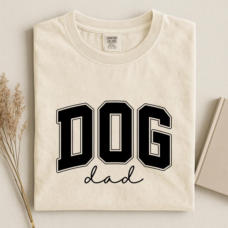 May include: Cream-colored t-shirt with the words "DOG dad" in bold black lettering. The word "DOG" is in a collegiate-style font, and "dad" is written in a cursive script. The shirt has a crew neck and short sleeves.