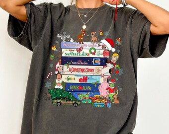 May include: A charcoal gray short-sleeve t-shirt featuring a graphic print of stacked VHS tapes labeled with classic holiday movie titles including The Santa Clause, Christmas Vacation, A Christmas Story, Elf, and Home Alone, surrounded by festive icons.