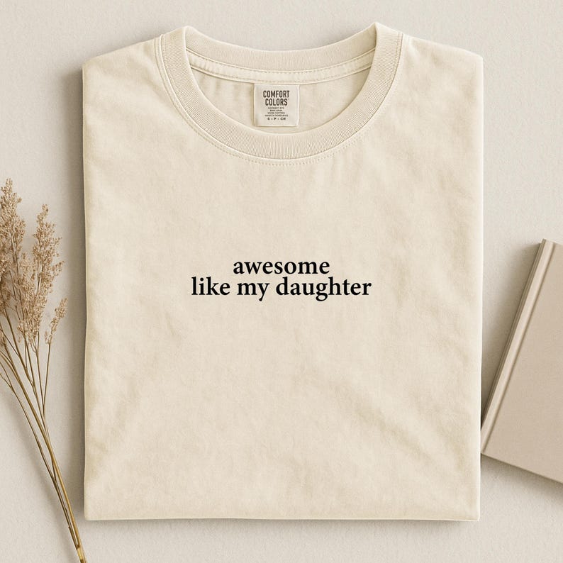 Puede incluir: Camiseta color crema con el texto negro "awesome like my daughter". La camiseta tiene cuello redondo y una etiqueta en el cuello. Un libro y plantas secas est&aacute;n en el fondo.