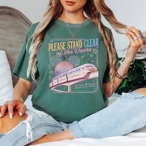 Puede incluir: Camiseta verde con el texto "PLEASE STAND CLEAR of the DOORS" sobre un gr&aacute;fico de un monorra&iacute;l, fuegos artificiales y una esfera. La camiseta se combina con vaqueros azul claro.