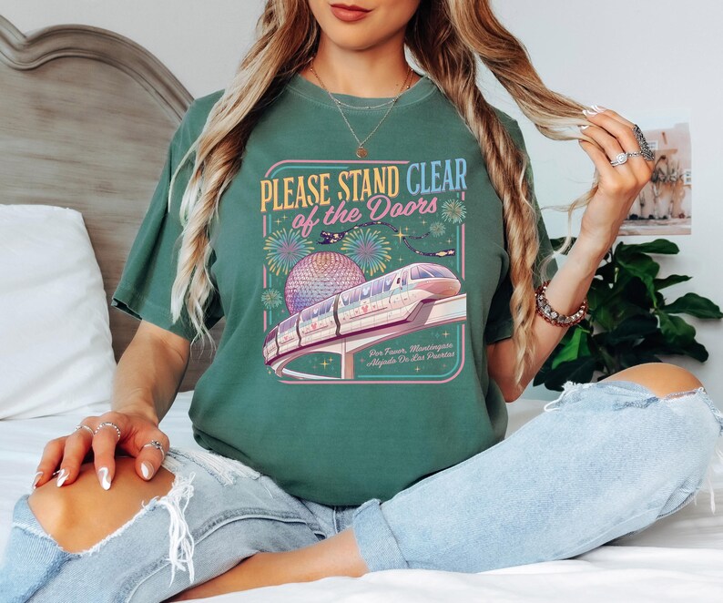 Puede incluir: Camiseta verde con el texto "PLEASE STAND CLEAR of the DOORS" sobre un gr&aacute;fico de un monorra&iacute;l, fuegos artificiales y una esfera. La camiseta se combina con vaqueros azul claro.