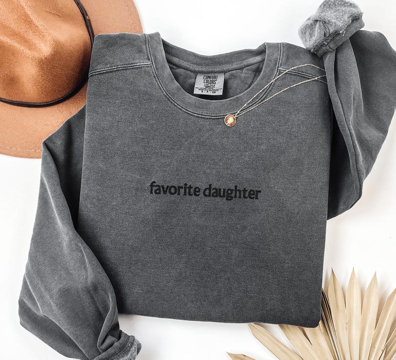 May include: A dark grey sweatshirt with the words "favorite daughter" printed in black. A gold pendant necklace is draped over the front. A brown hat is visible in the upper left corner.