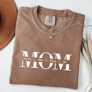 May include: A brown Comfort Colors t-shirt with the word "MOM" in large white letters. Below the word "MOM" are the names Amelia, Alex, Mina, and Lila. A silver necklace with a small pendant is on the shirt.