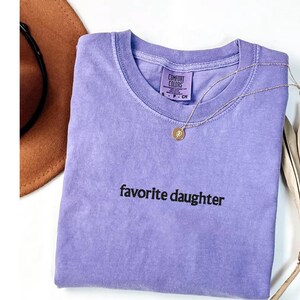 May include: A lilac Comfort Colors t-shirt with the words "favorite daughter" in black. A gold necklace with a pendant lies on the shirt. A brown hat is partially visible in the upper left corner.