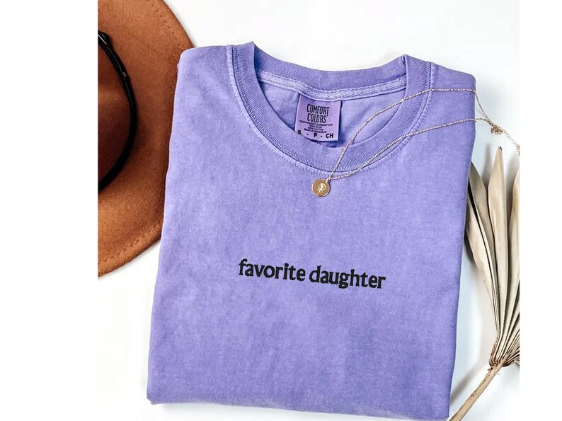 May include: A lilac Comfort Colors t-shirt with the words "favorite daughter" in black. A gold necklace with a pendant lies on the shirt. A brown hat is partially visible in the upper left corner.