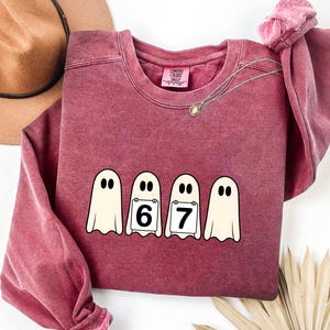 May include: A maroon crewneck sweatshirt featuring a graphic of four cartoon ghosts. Two ghosts hold signs with the numbers 6 and 7. The sweatshirt has long sleeves and a Comfort Colors tag.