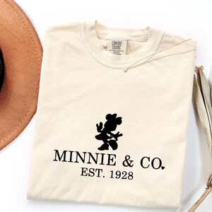 Pu&ograve; includere: T-shirt color crema con silhouette nera di Minnie Mouse e la scritta "MINNIE & CO. EST. 1928." Un cappello marrone e una pianta secca sono visibili sullo sfondo. La maglietta ha un'etichetta Comfort Colors.