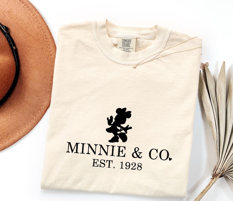 Pu&ograve; includere: T-shirt color crema con silhouette nera di Minnie Mouse e la scritta "MINNIE & CO. EST. 1928." Un cappello marrone e una pianta secca sono visibili sullo sfondo. La maglietta ha un'etichetta Comfort Colors.