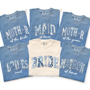 May include: A collection of blue and cream-colored t-shirts with text for a wedding party. The blue shirts say "Mother of the Bride," "Maid of Honor," "Mother of the Groom," "Bridesmaid," and "Matron of Honor." The cream shirt says "Bride."