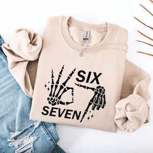 May include: A beige sweatshirt with the words "SIX SEVEN!" in black, accompanied by a graphic of two skeleton hands. A silver necklace with a small pendant is on the neckline.