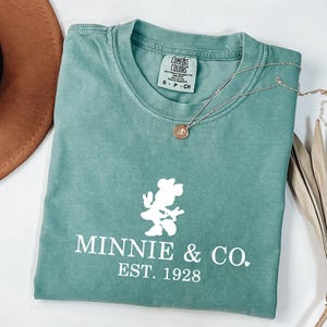 Pu&ograve; includere: Una maglietta verde acqua con una grafica bianca di Minnie Mouse e la scritta "MINNIE & CO. EST. 1928". Una collana d'oro con un piccolo ciondolo &egrave; sulla maglietta. Un cappello marrone e foglie di palma secche sono sullo sfondo.