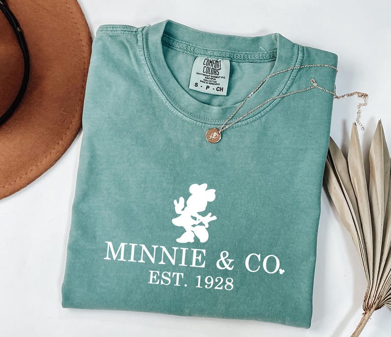 Pu&ograve; includere: Una maglietta verde acqua con una grafica bianca di Minnie Mouse e la scritta "MINNIE & CO. EST. 1928". Una collana d'oro con un piccolo ciondolo &egrave; sulla maglietta. Un cappello marrone e foglie di palma secche sono sullo sfondo.