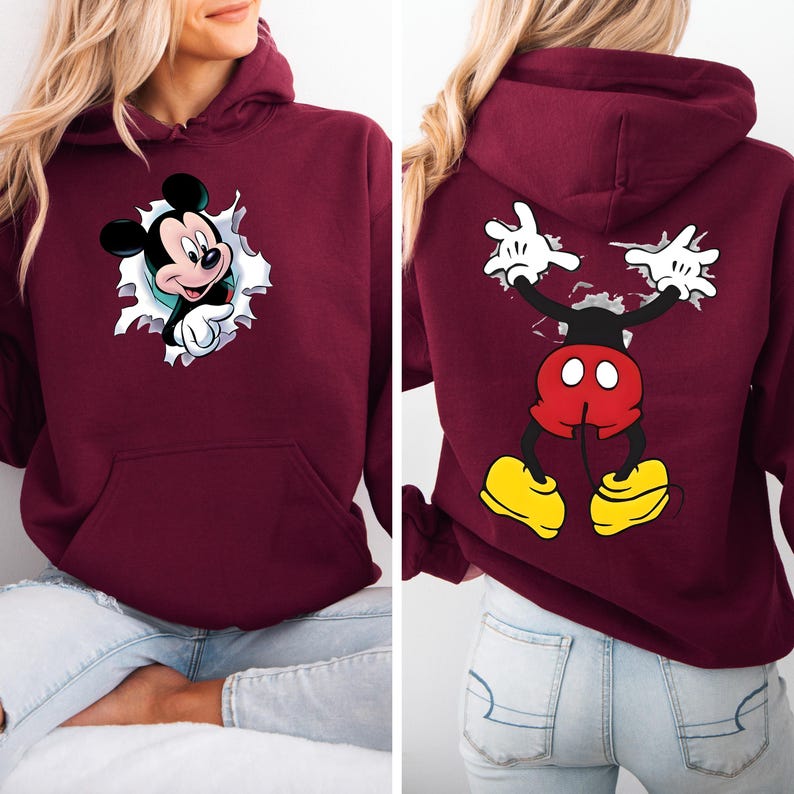 May include: Burgundy hooded sweatshirt featuring Mickey Mouse. The front shows Mickey breaking through a white-edged hole, while the back displays Mickey's arms and legs. The design includes black, red, yellow, and white details.