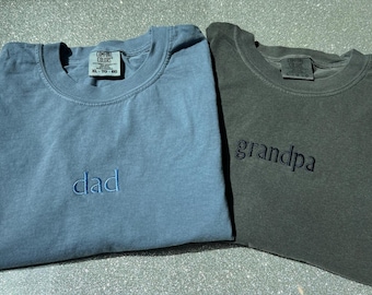 Puede incluir: Dos camisetas de cuello redondo con etiquetas Comfort Colors. La camiseta de la izquierda es azul pizarra con la palabra "dad" bordada en hilo azul claro. La de la derecha es gris carbón con la palabra "grandpa" bordada en hilo negro.