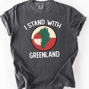 Pu&ograve; includere: T-shirt grigio scuro con la scritta bianca "I STAND WITH GREENLAND". La maglietta presenta una grafica circolare con una mappa della Groenlandia in verde, su sfondo rosso e bianco.