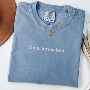K&ouml;nnte beinhalten: Blaues Comfort Colors T-Shirt mit dem wei&szlig;en Aufdruck "favorite custom". Eine goldene Halskette mit Anh&auml;nger liegt auf dem Shirt. Ein brauner Hut und eine getrocknete Pflanze sind im Hintergrund.