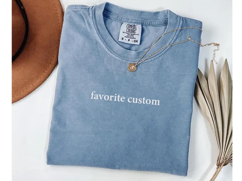 K&ouml;nnte beinhalten: Blaues Comfort Colors T-Shirt mit dem wei&szlig;en Aufdruck "favorite custom". Eine goldene Halskette mit Anh&auml;nger liegt auf dem Shirt. Ein brauner Hut und eine getrocknete Pflanze sind im Hintergrund.