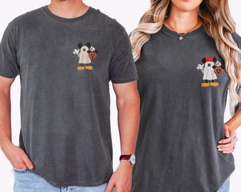 May include: Two charcoal gray crew neck t-shirts featuring embroidered designs on the left chest. One shirt displays a ghost character wearing mouse ears and holding a pumpkin bucket. The other shirt shows a ghost with a red bow and pumpkin bucket.