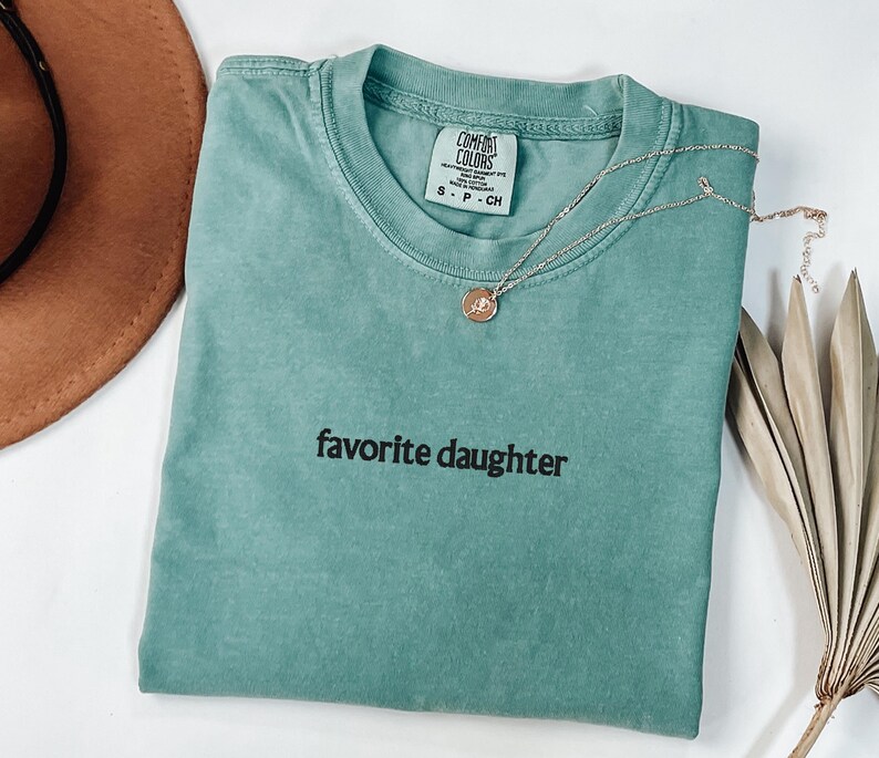 May include: A teal Comfort Colors t-shirt with the words "favorite daughter" in black. A gold necklace with a small pendant is on the shirt. A brown hat and dried palm leaves are in the background.