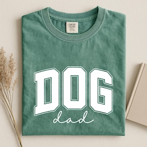 May include: A sage green t-shirt with the words "DOG dad" printed in white. The word "DOG" is in a bold, block letter font, and "dad" is in a cursive font. The shirt is folded neatly.