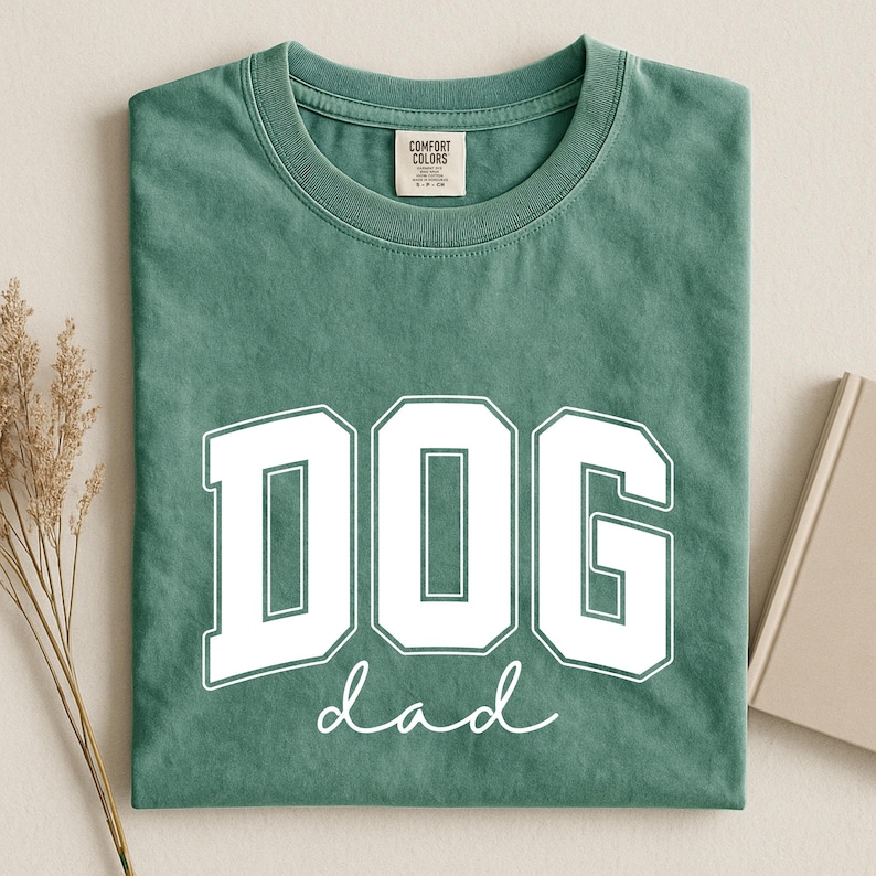 May include: A sage green t-shirt with the words "DOG dad" printed in white. The word "DOG" is in a bold, block letter font, and "dad" is in a cursive font. The shirt is folded neatly.