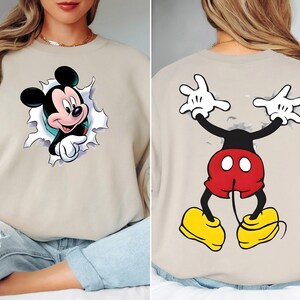 May include: Beige sweatshirt featuring a Mickey Mouse graphic. The front shows Mickey breaking through a torn background, while the back displays Mickey's arms and legs. Mickey is wearing red shorts, yellow shoes, and white gloves. A fun, cartoon-themed apparel item.