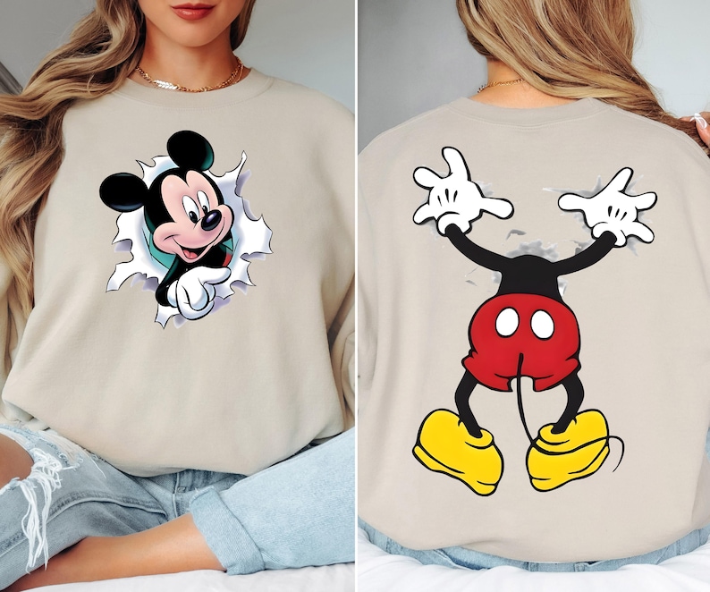 May include: Beige sweatshirt featuring a Mickey Mouse graphic. The front shows Mickey breaking through a torn background, while the back displays Mickey's arms and legs. Mickey is wearing red shorts, yellow shoes, and white gloves. A fun, cartoon-themed apparel item.