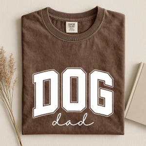 May include: A brown t-shirt with the words "DOG dad" printed in white. The word "DOG" is in a bold, block letter font, and "dad" is in a cursive font. The shirt has a crew neck and a tag that says "Comfort Colors".