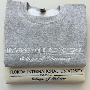 May include: Two folded sweatshirts, one gray and one cream-colored. The gray sweatshirt displays "UNIVERSITY OF ILLINOIS CHICAGO" and "College of Pharmacy." The cream sweatshirt shows "FLORIDA INTERNATIONAL UNIVERSITY" and "College of Medicine."