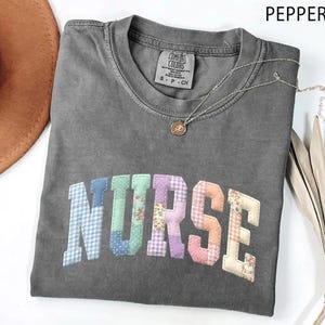 May include: A gray Comfort Colors t-shirt with the word "NURSE" in patchwork letters. The letters are various colors and patterns. A silver necklace with a pendant is on the shirt. A brown hat is in the upper left corner.
