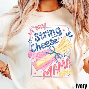 May include: Ivory-colored t-shirt with a graphic design. The design features the text "In my String Cheese Mama Era" in blue and pink, along with illustrations of string cheese and a pink wrapper.