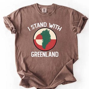 Pu&ograve; includere: T-shirt marrone con la scritta bianca "I STAND WITH GREENLAND". La maglietta presenta una grafica circolare con una mappa verde della Groenlandia su sfondo bianco, rosso e beige. Le maniche sono arrotolate.