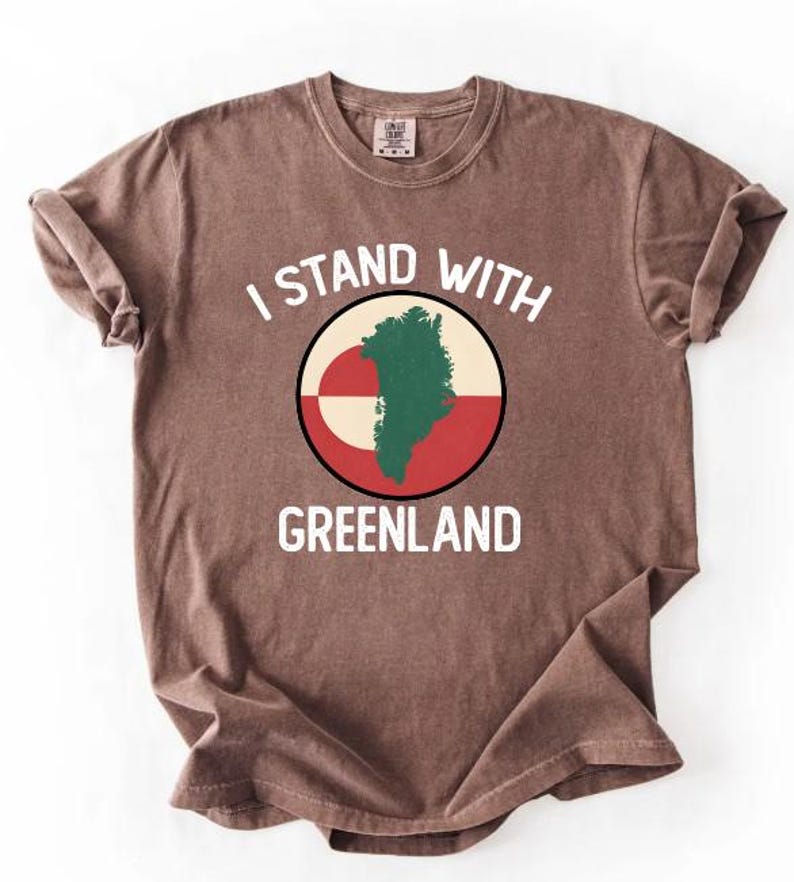 Pu&ograve; includere: T-shirt marrone con la scritta bianca "I STAND WITH GREENLAND". La maglietta presenta una grafica circolare con una mappa verde della Groenlandia su sfondo bianco, rosso e beige. Le maniche sono arrotolate.