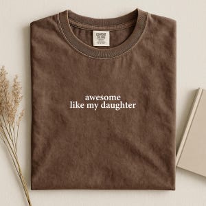 May include: A brown t-shirt with the text "awesome like my daughter" in white. The shirt has a crew neck and a tag that says "Comfort Colors". The shirt is folded and placed on a neutral surface.