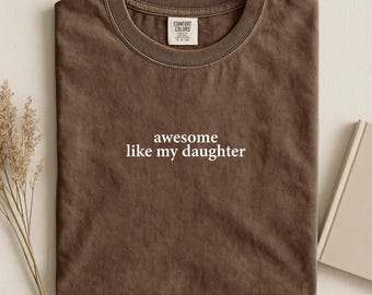 May include: A brown t-shirt with the text "awesome like my daughter" in white. The shirt has a crew neck and a tag that says "Comfort Colors". The shirt is folded and placed on a neutral surface.