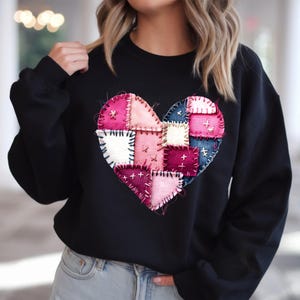 May include: Black sweatshirt featuring a large, patchwork heart design. The heart is composed of various fabric squares in shades of pink, white, and blue, stitched together with visible thread. A casual, comfortable style.