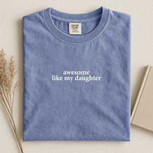 Puede incluir: Una camiseta azul Comfort Colors con el texto blanco "awesome like my daughter". La camiseta est&aacute; doblada y descansa sobre una superficie neutra. Una peque&ntilde;a etiqueta es visible cerca del cuello.
