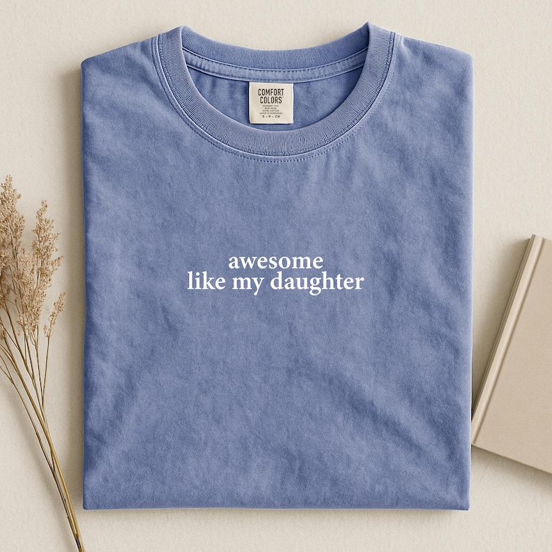 Puede incluir: Una camiseta azul Comfort Colors con el texto blanco "awesome like my daughter". La camiseta est&aacute; doblada y descansa sobre una superficie neutra. Una peque&ntilde;a etiqueta es visible cerca del cuello.