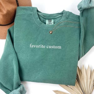 K&ouml;nnte beinhalten: Ein teal-farbenes Sweatshirt mit den Worten "favorite custom" in wei&szlig;er Schrift. Das Sweatshirt hat einen Rundhalsausschnitt und lange &Auml;rmel mit gerollten Manschetten. Eine goldene Halskette mit Anh&auml;nger ist &uuml;ber die Vorderseite drapiert.