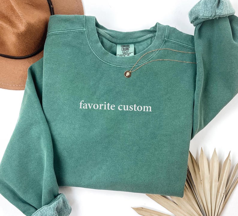 K&ouml;nnte beinhalten: Ein teal-farbenes Sweatshirt mit den Worten "favorite custom" in wei&szlig;er Schrift. Das Sweatshirt hat einen Rundhalsausschnitt und lange &Auml;rmel mit gerollten Manschetten. Eine goldene Halskette mit Anh&auml;nger ist &uuml;ber die Vorderseite drapiert.