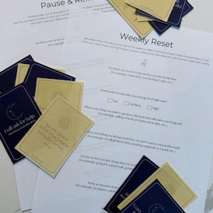 May include: A collection of affirmation cards with navy blue and tan designs, scattered on top of printed worksheets. The cards feature sun and moon illustrations and phrases like "This is really hard, and I'm doing it."