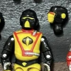 May include: A yellow and black action figure with a helmet and mask. The figure has a red and yellow vest with a cobra emblem. The figure's arms and legs are black and yellow. The figure is a vintage toy.