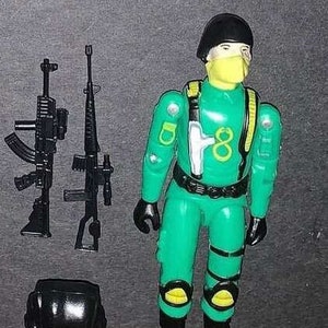 May include: A green action figure with a black helmet and a yellow face mask. The figure is holding two black guns and has a yellow number "8" on its chest.