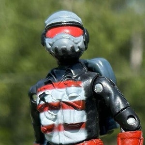Puede incluir: Una figura de acción negra y roja con un casco gris y una visera roja. La figura lleva una camisa a rayas blancas y negras con un cinturón rojo y pantalones negros. La figura sostiene un rifle gris.