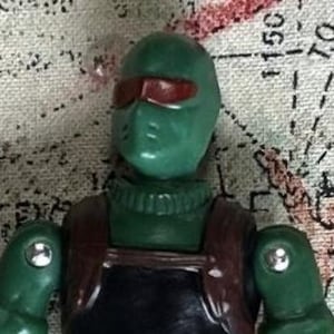 May include: A green action figure with a red visor and a brown backpack. The figure is wearing a green jumpsuit and has a black belt.